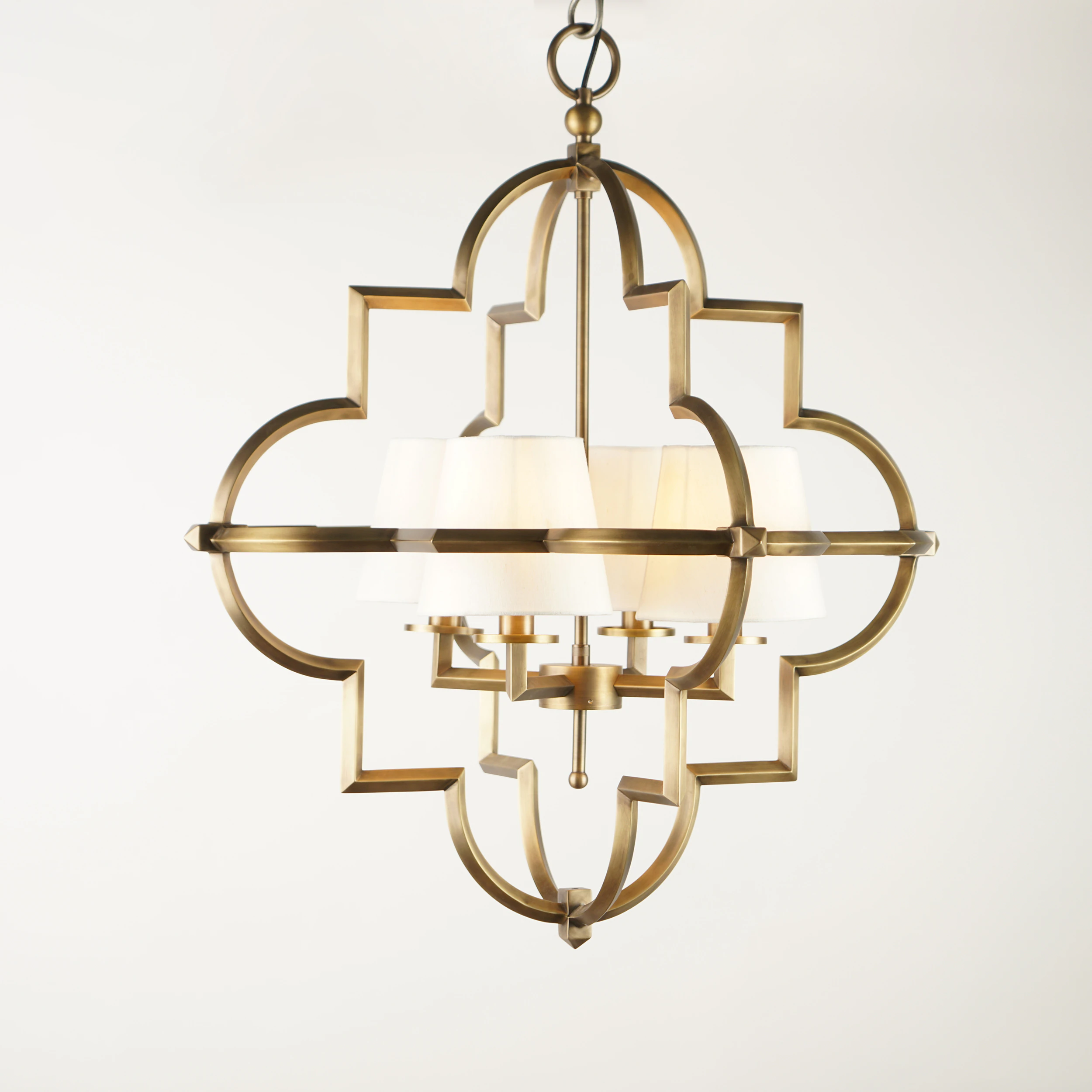 The QUATREFOIL PENDANT is a modern brass chandelier featuring a quatrefoil-shaped frame and six white fabric lampshades, shown suspended against a plain white background. The QUATREFOIL PENDANT is a modern brass chandelier featuring a quatrefoil-shaped frame and six white fabric lampshades, shown suspended against a plain white background.