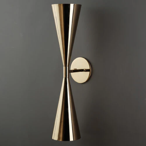 The SELENE WALL SCONCE features two cone-shaped brass shades joined at the tips and mounted on a round base, shown against a dark gray wall.