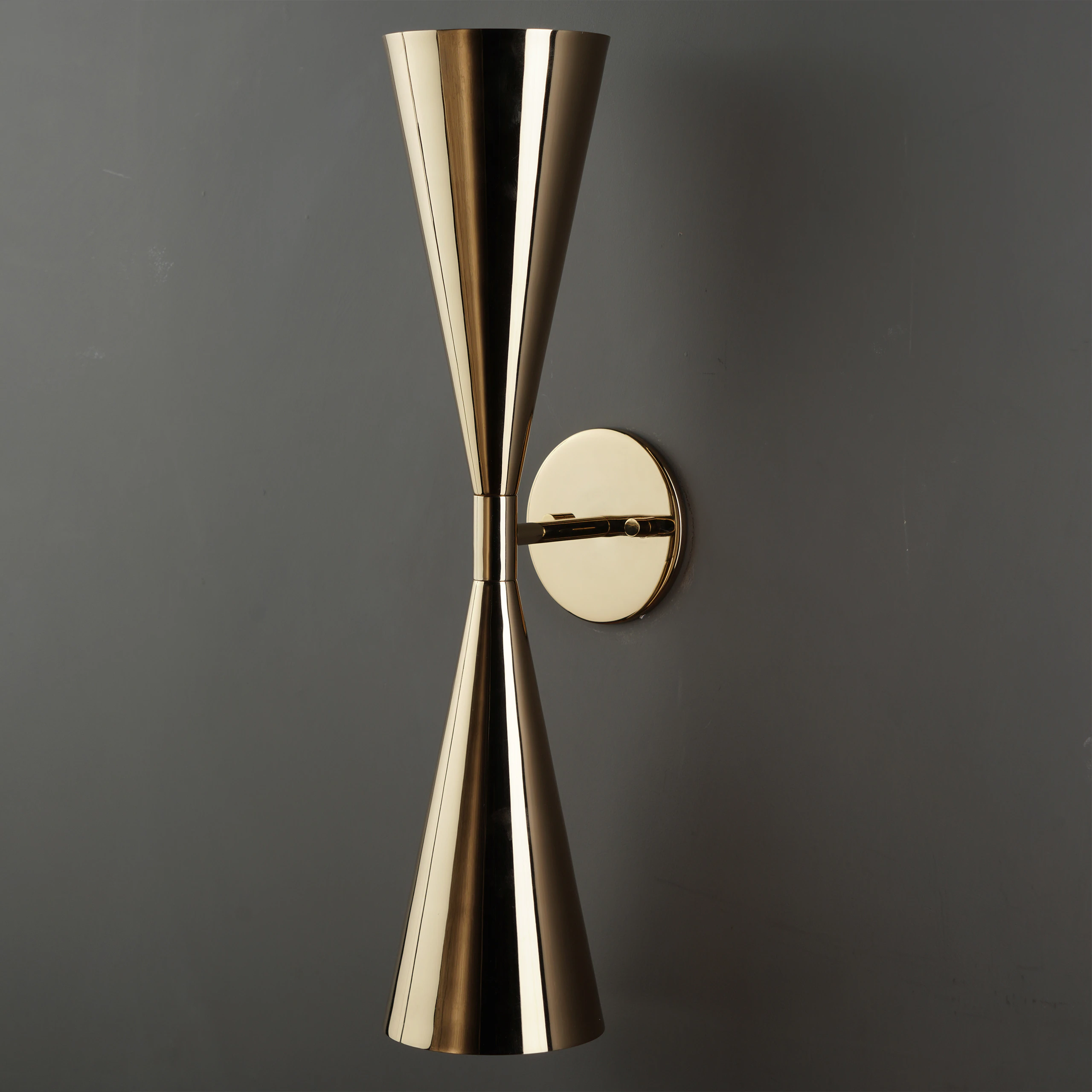 The SELENE WALL SCONCE features two cone-shaped brass shades joined at the tips and mounted on a round base, shown against a dark gray wall. The SELENE WALL SCONCE features two cone-shaped brass shades joined at the tips and mounted on a round base, shown against a dark gray wall.