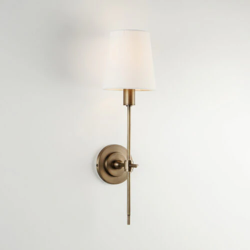 The BROOKLYN WALL SCONCE features a round brass base with a slender vertical arm and a white tapered lampshade, mounted on a plain white wall.