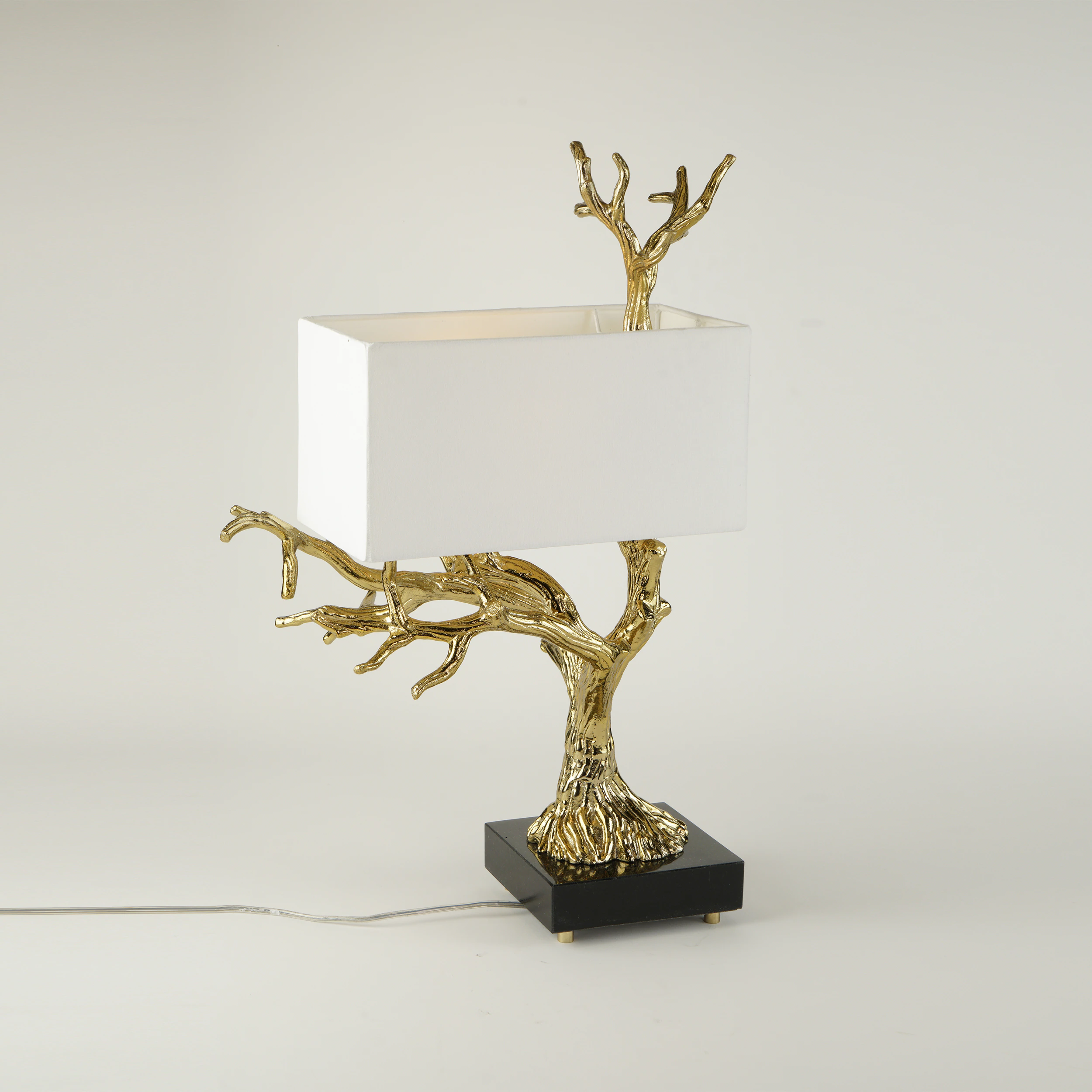 The ALBERO TABLE LAMP features a gold tree-branch-shaped base, a rectangular white lampshade, and sits on a black square platform with an electrical cord. The ALBERO TABLE LAMP features a gold tree-branch-shaped base, a rectangular white lampshade, and sits on a black square platform with an electrical cord.