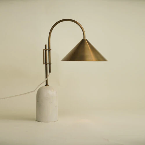 The BROOKS TABLE LAMP features a white marble base, a curved brass stem, and a conical brass shade. Shown illuminated against an off-white background, it casts light downward in a modern style.