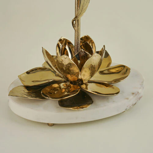 The MAGNOLIA TABLE LAMP features a golden metal lotus flower with detailed petals, set on a round white marble base, displayed against a simple light background.