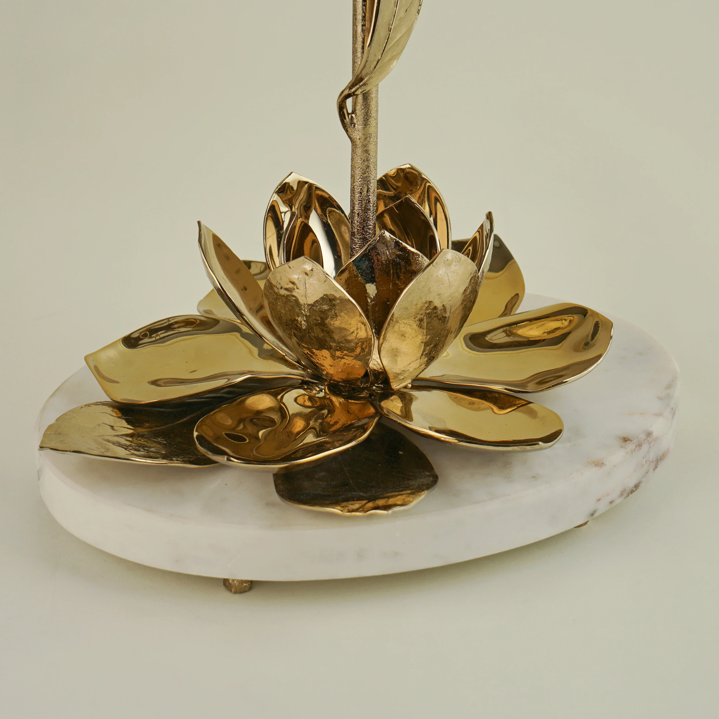 The MAGNOLIA TABLE LAMP features a golden metal lotus flower with detailed petals, set on a round white marble base, displayed against a simple light background. The MAGNOLIA TABLE LAMP features a golden metal lotus flower with detailed petals, set on a round white marble base, displayed against a simple light background.