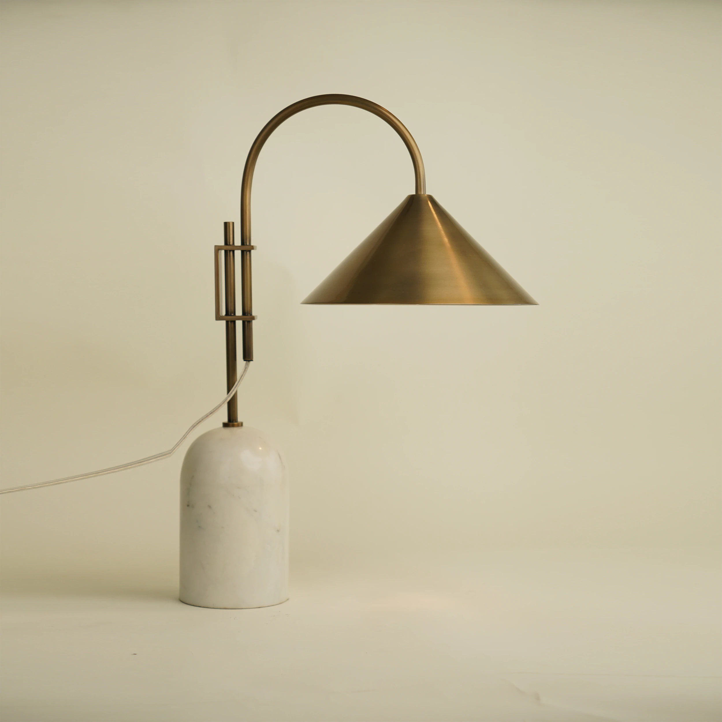 The BROOKS TABLE LAMP features a white marble base, a curved brass stem, and a conical brass shade. Shown illuminated against an off-white background, it casts light downward in a modern style. The BROOKS TABLE LAMP features a white marble base, a curved brass stem, and a conical brass shade. Shown illuminated against an off-white background, it casts light downward in a modern style.