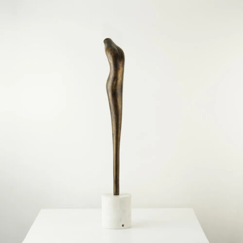 The VEGA SCULPTURE, a slender and elongated bronze piece with a subtle curve, is mounted on a round white marble base and displayed on a white surface against a plain white background.