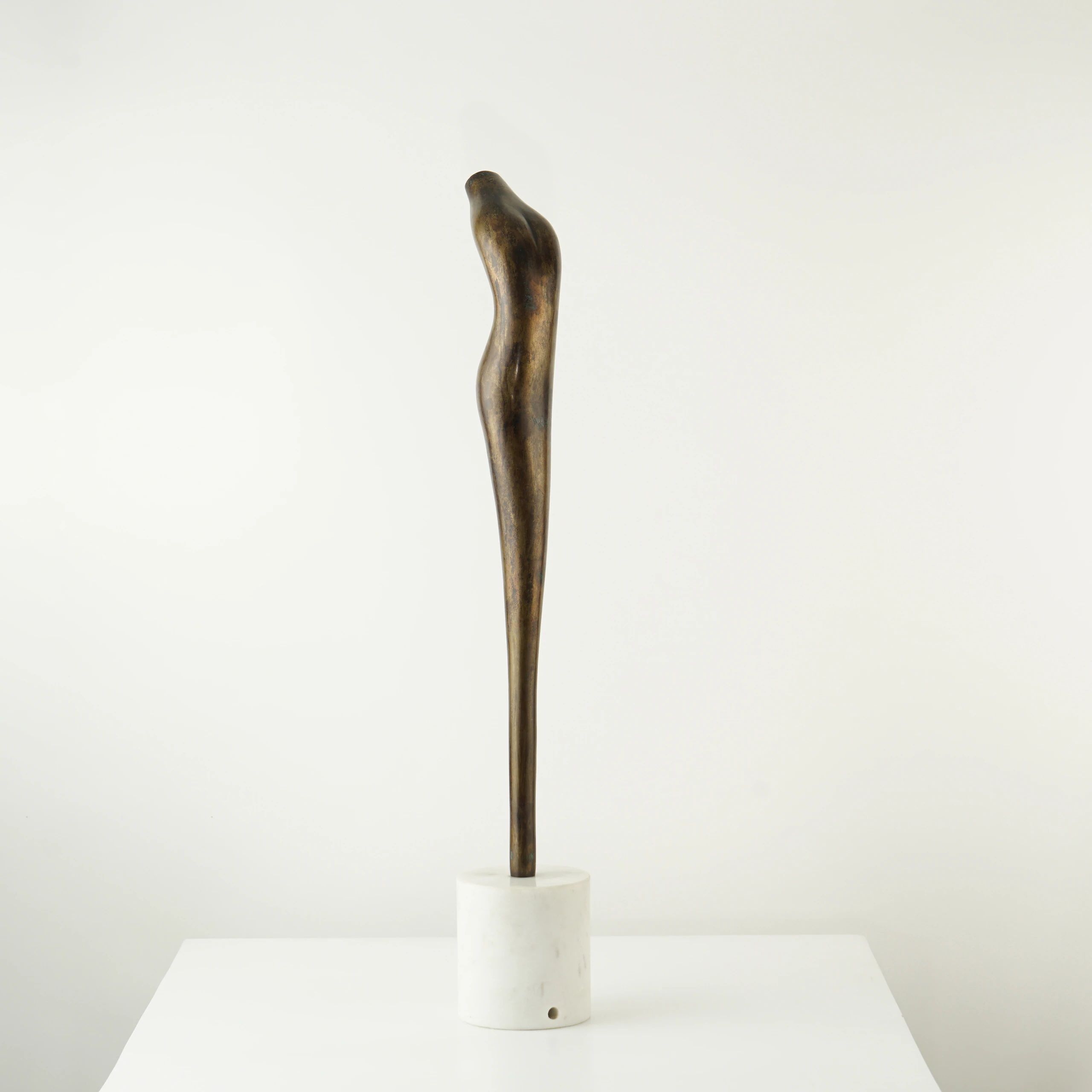 The VEGA SCULPTURE, a slender and elongated bronze piece with a subtle curve, is mounted on a round white marble base and displayed on a white surface against a plain white background. The VEGA SCULPTURE, a slender and elongated bronze piece with a subtle curve, is mounted on a round white marble base and displayed on a white surface against a plain white background.
