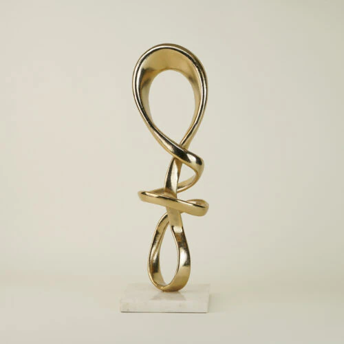 The ALTAIR SCULPTURE features a modern abstract gold design with intertwined loops, standing vertically on a white marble base against a plain light background.