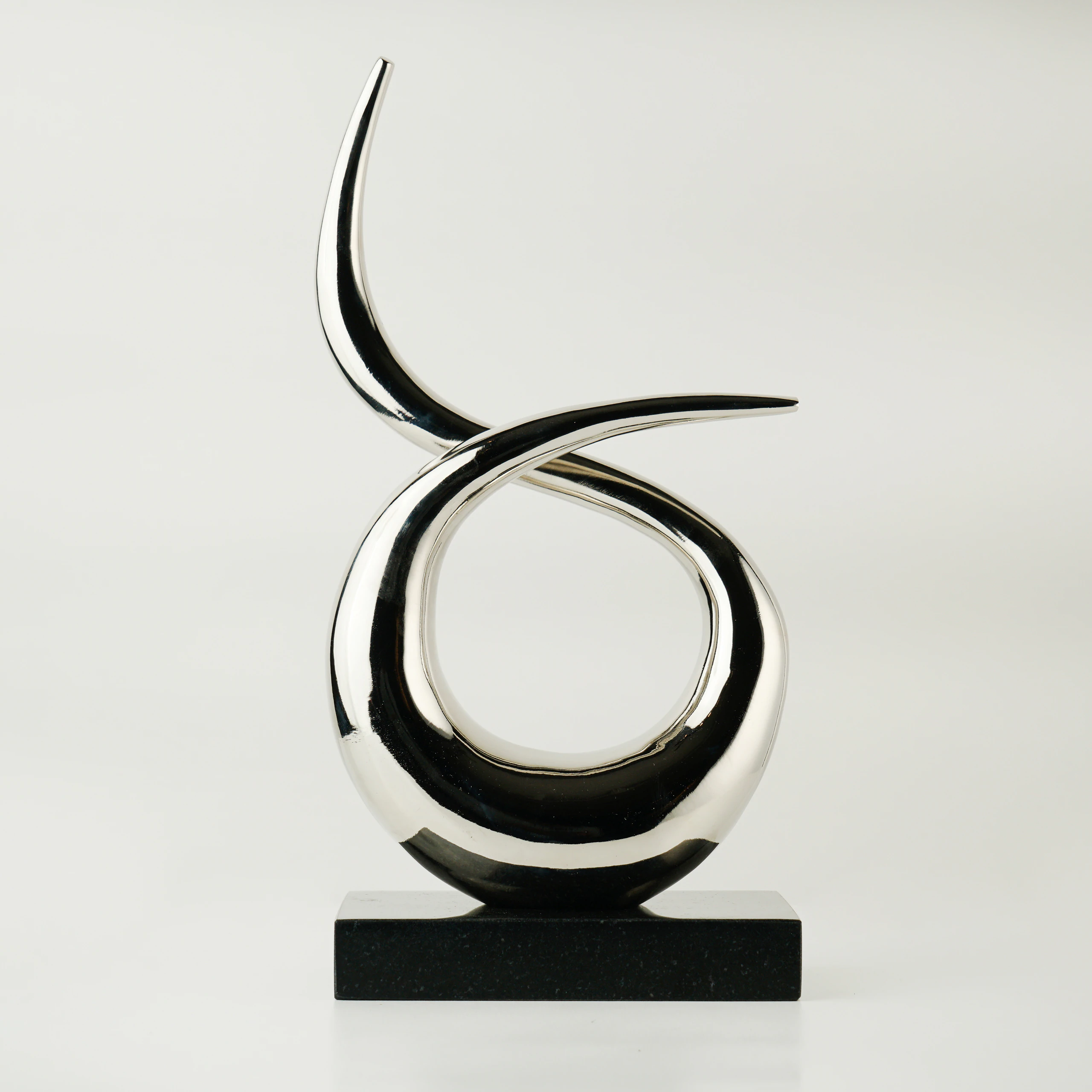 The VEGA SCULPTURE features two polished, curved metallic forms intertwined above a rectangular black base, displayed against a plain white background. The VEGA SCULPTURE features two polished, curved metallic forms intertwined above a rectangular black base, displayed against a plain white background.