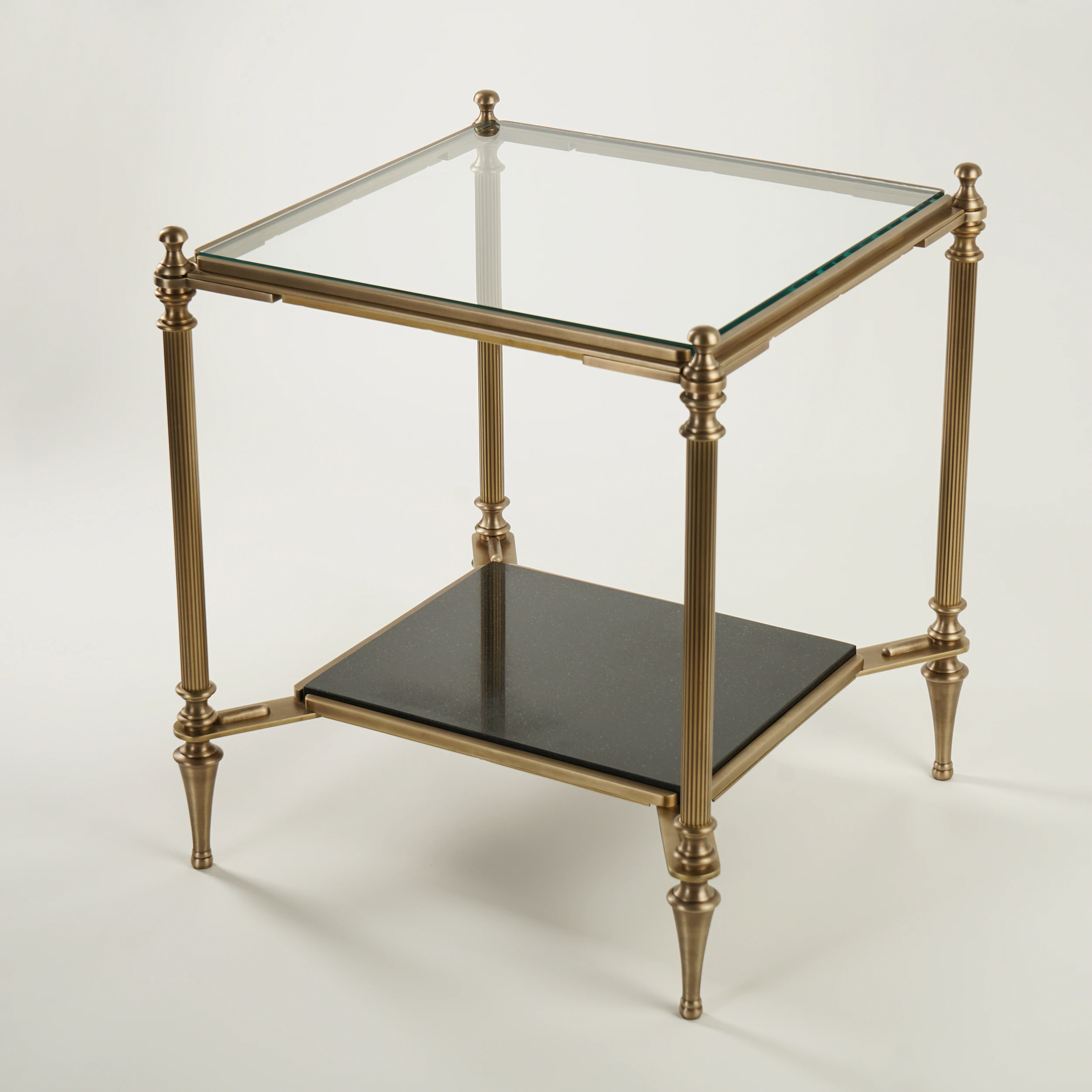 The MEDFORD END TABLE features a square glass top, gold metal frame, four tapered legs, and a lower black shelf, displayed against a plain white background. The MEDFORD END TABLE features a square glass top, gold metal frame, four tapered legs, and a lower black shelf, displayed against a plain white background.