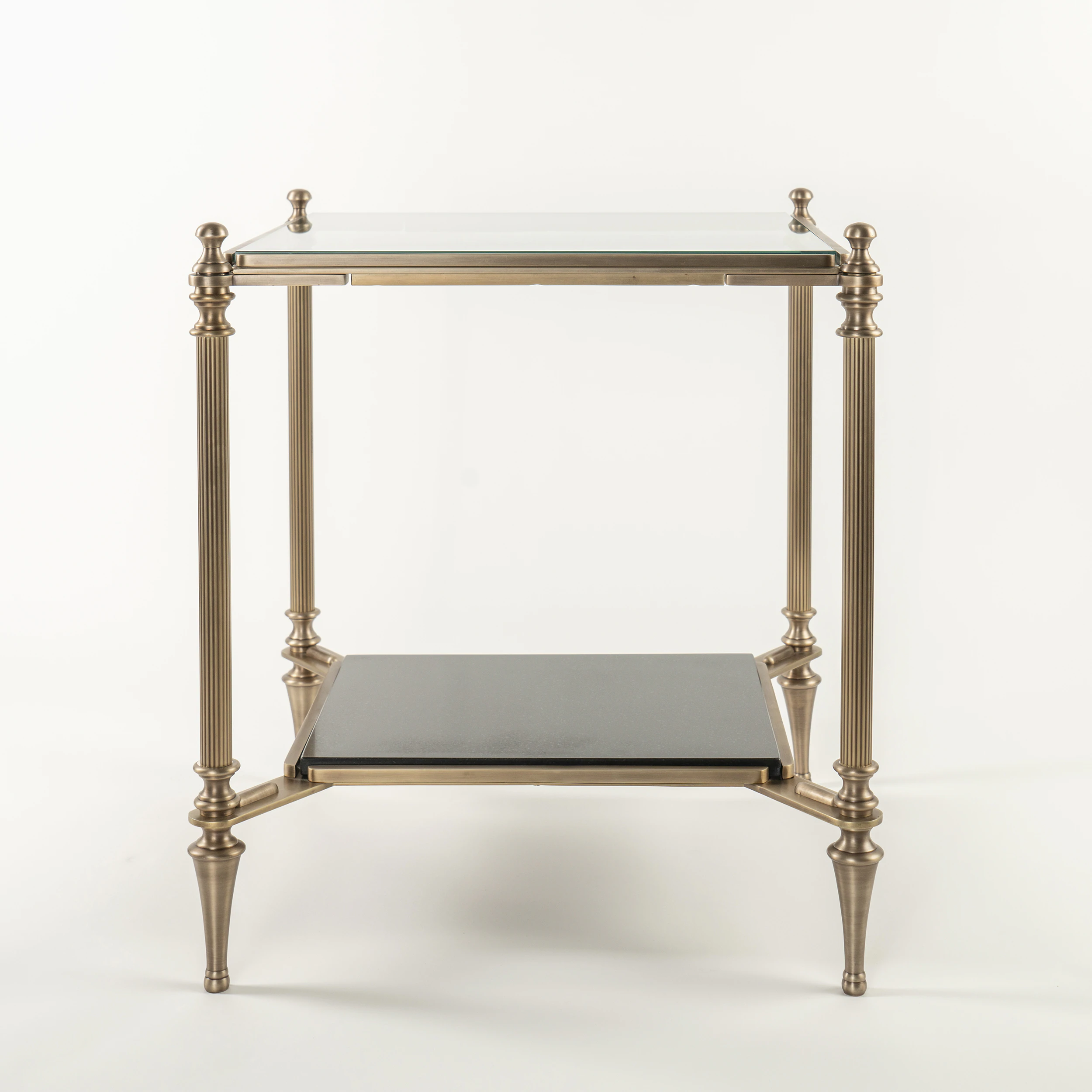 The MEDFORD END TABLE features a glass top, gold-toned metal frame, four slender fluted legs, and a black lower shelf in an elegant, minimalist design. Shown against a plain white background. The MEDFORD END TABLE features a glass top, gold-toned metal frame, four slender fluted legs, and a black lower shelf in an elegant, minimalist design. Shown against a plain white background.