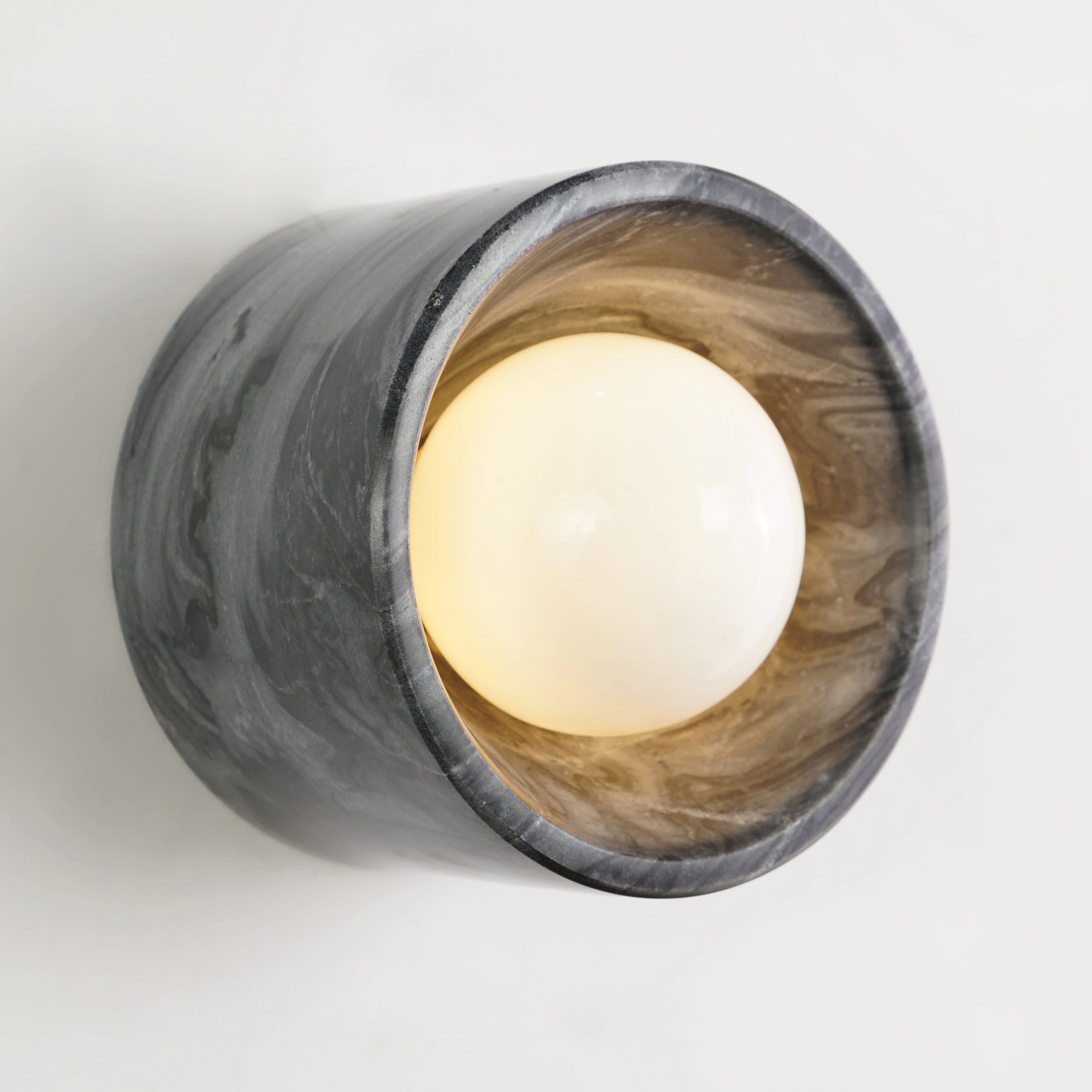 The NANO STONE WALL SCONCE features a gray marbled stone outer shell and a glowing white bulb at its center, mounted on a white wall. The NANO STONE WALL SCONCE features a gray marbled stone outer shell and a glowing white bulb at its center, mounted on a white wall.