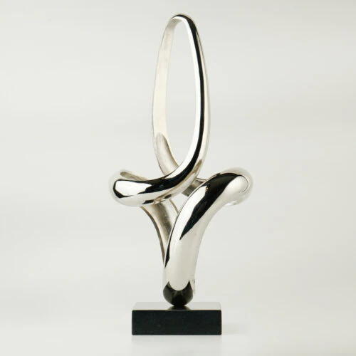 The VEGA SCULPTURE is a polished abstract silver metal piece with smooth, curved loops, standing vertically on a small black rectangular base. Set against a plain white background, its reflective surface is highlighted.